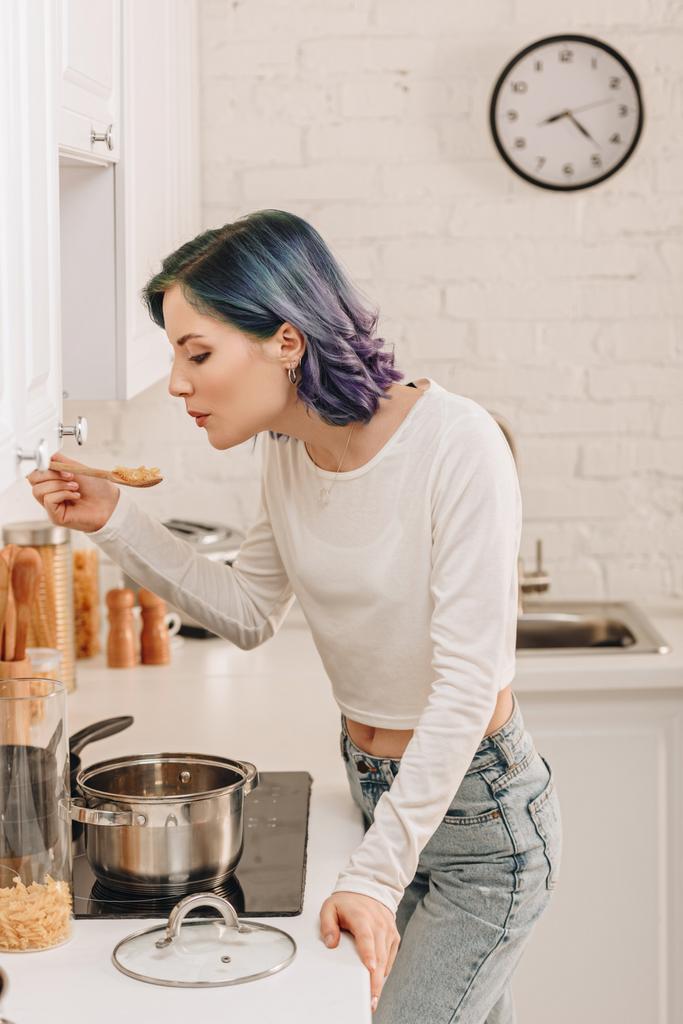 Dívka s barevnými vlasy foukání na stěrku s těstovinami v blízkosti kuchyňského sporáku - Fotografie, Obrázek