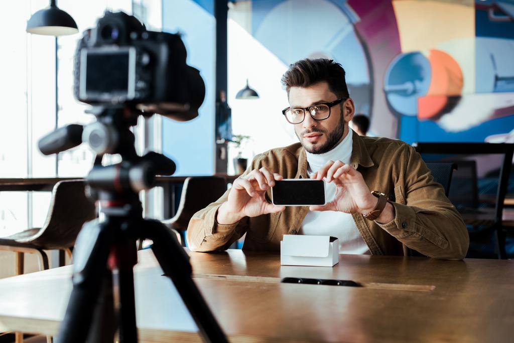 Enfoque selectivo del bloguero frente a la cámara digital mostrando el teléfono inteligente en la mesa en el espacio de coworking
 - Foto, Imagen