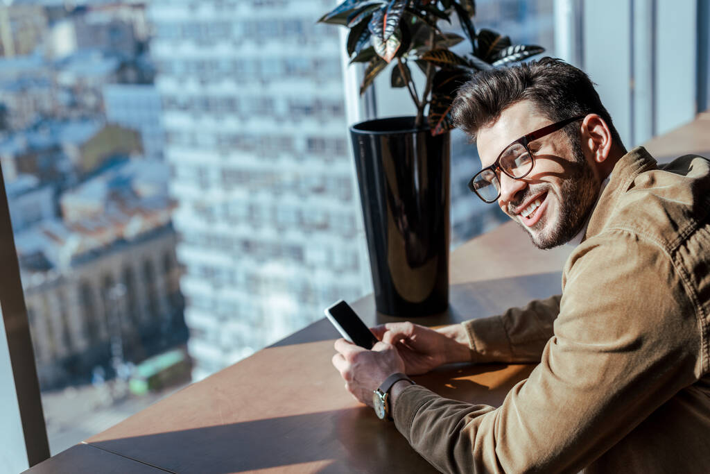 Vista de ángulo alto del trabajador de TI con teléfono inteligente sonriendo en la mesa con maceta cerca de la ventana
 - Foto, imagen