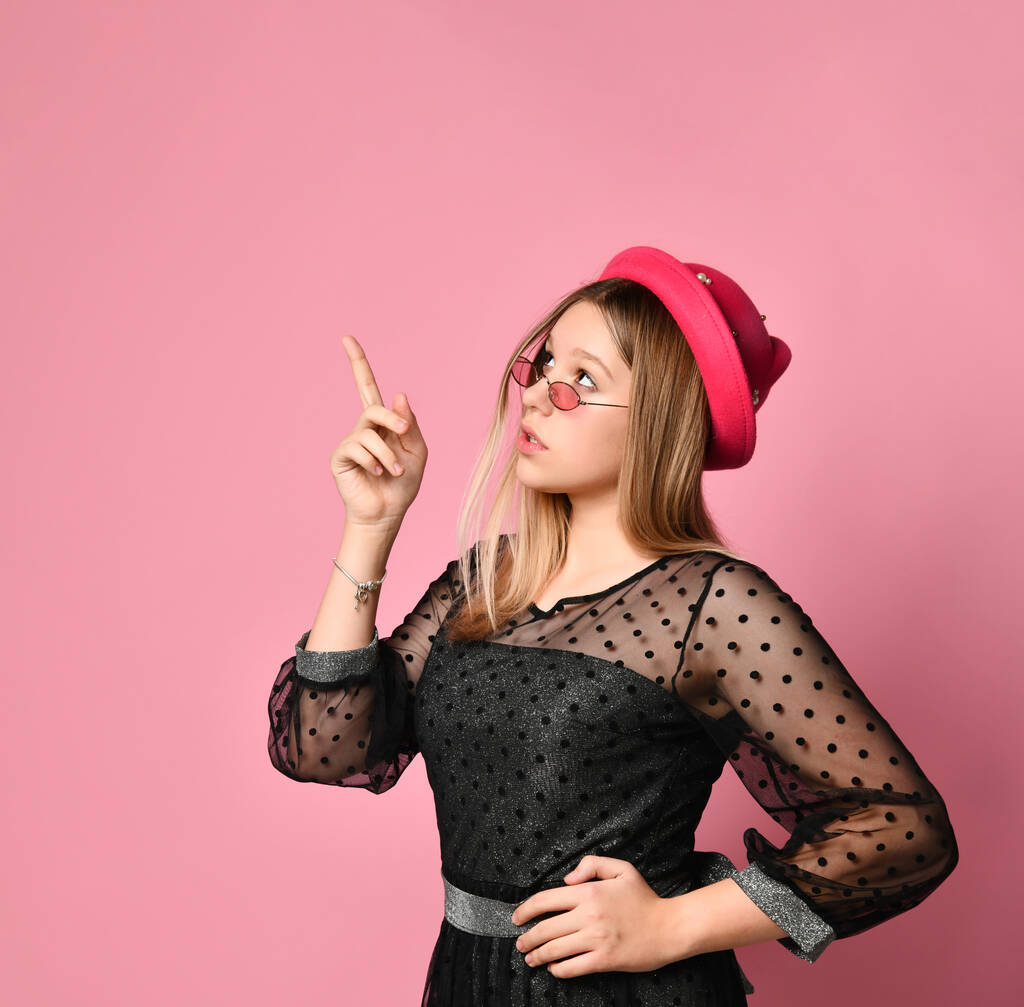 Chica adolescente en gafas de sol, vestido negro y sombrero rojo. Señala algo con el dedo índice, posando sobre un fondo rosa. De cerca.
 - Foto, Imagen