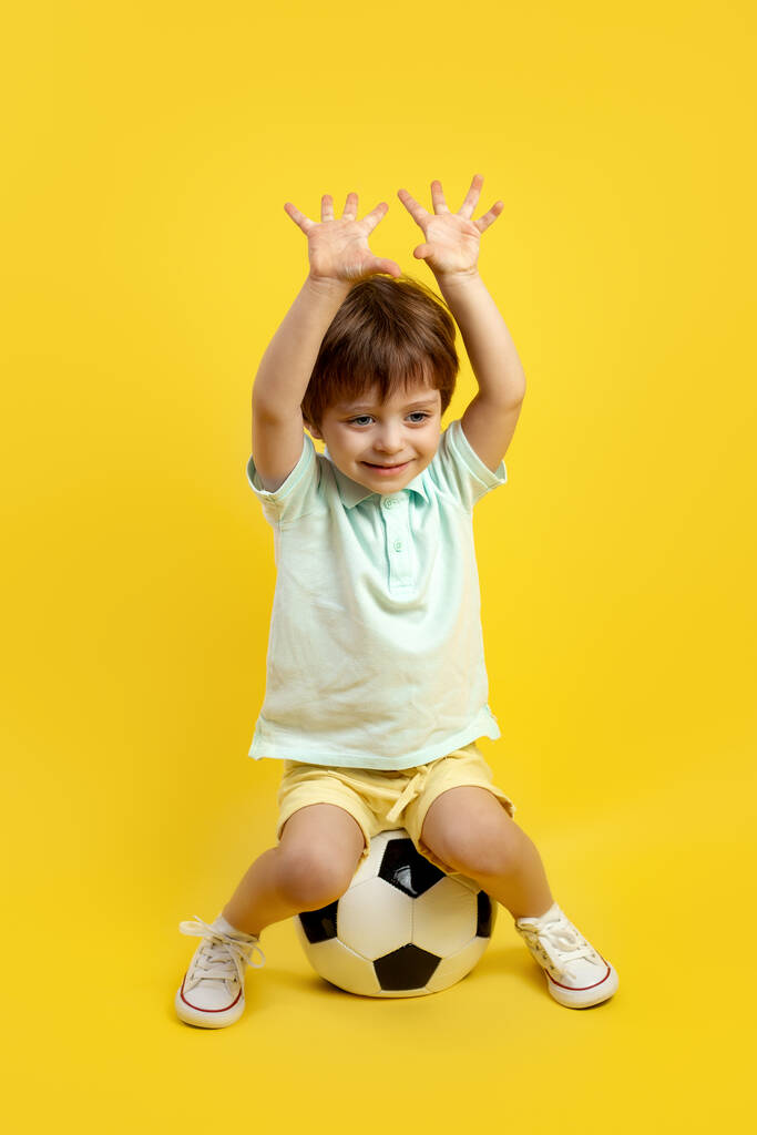Grappig jongetje in casual zomer outfit zitten op de voetbal en poseren met handen omhoog over gele achtergrond. - Foto, afbeelding