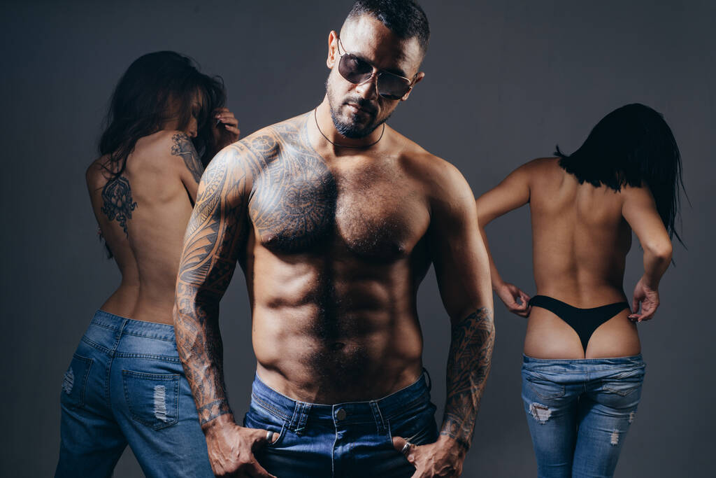 Sexy muscular macho man in sunglasses with two hot girls studio photo. Women with sexy fit ass standing behind handsome tattooed man with sexy torso. - Zdjęcie, obraz