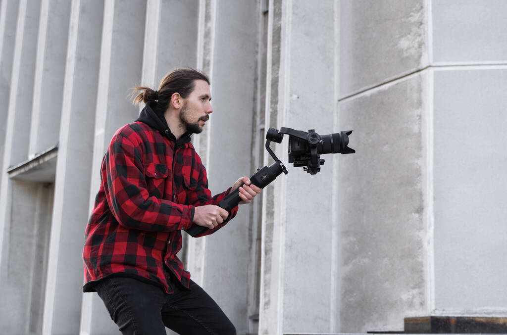 Young Professional videographer holding professional camera on 3-axis gimbal stabilizer. Pro equipment helps to make high quality video without shaking. Cameraman wearing red shirt making a videos. - Photo, Image