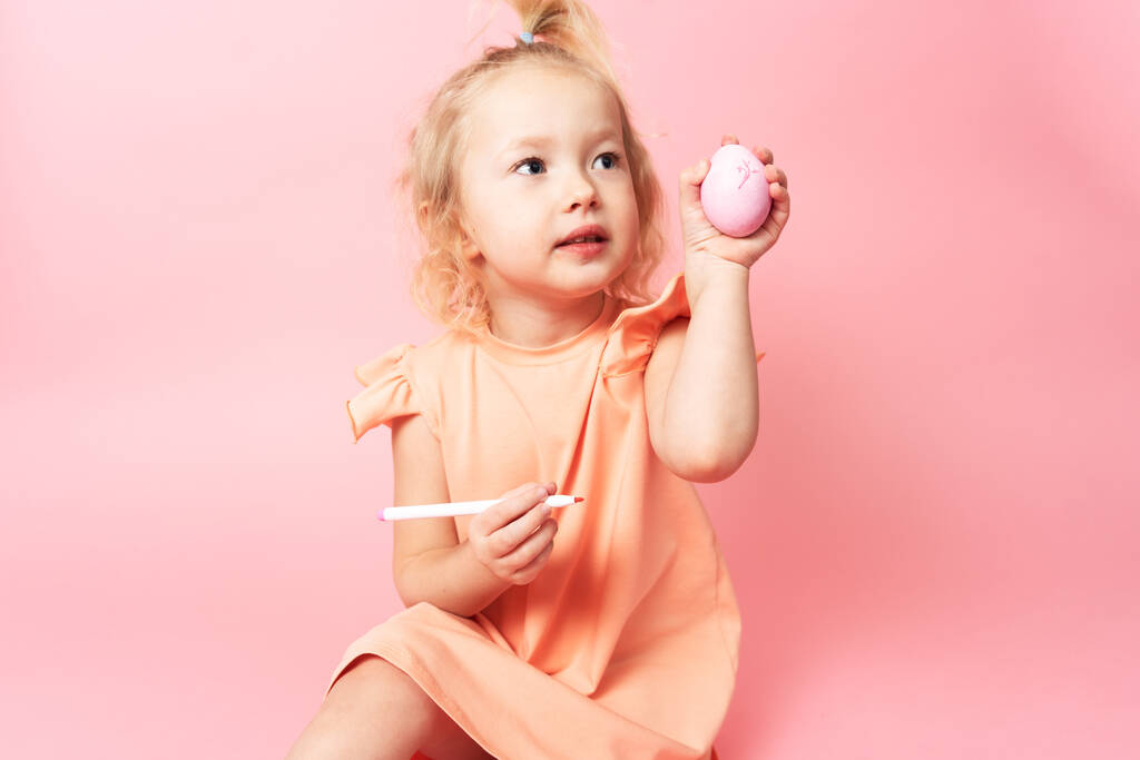 Καλό Πάσχα: ένα κοριτσάκι με φόρεμα κρατά ένα αυγό στο χέρι της και το ζωγραφίζει. Πορτρέτο σε ροζ φόντο. - Φωτογραφία, εικόνα