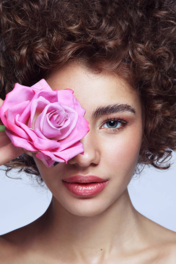 Retrato de cerca de la joven hermosa chica con el pelo rizado, maquillaje limpio y rosa en la mano
 - Foto, Imagen