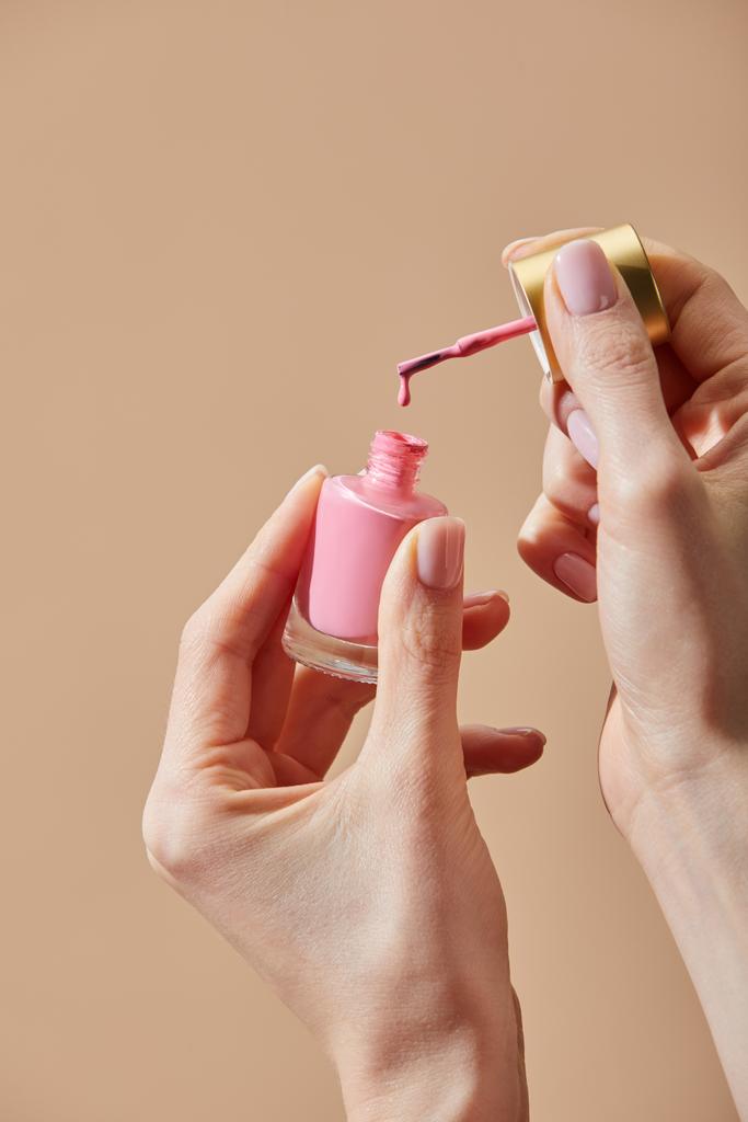 Обрізаний вид жінки, що тримає відкриту пляшку рожевого лаку для нігтів ізольовано на бежевому
 - Фото, зображення