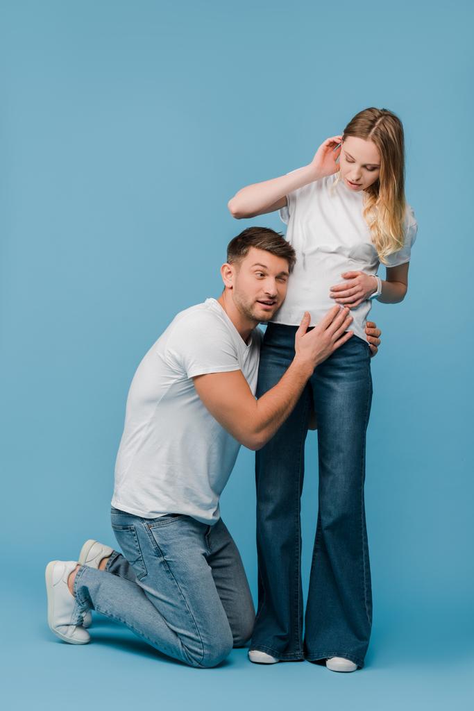 emotionele man luisteren buik van zwangere vrouw op blauw - Foto, afbeelding