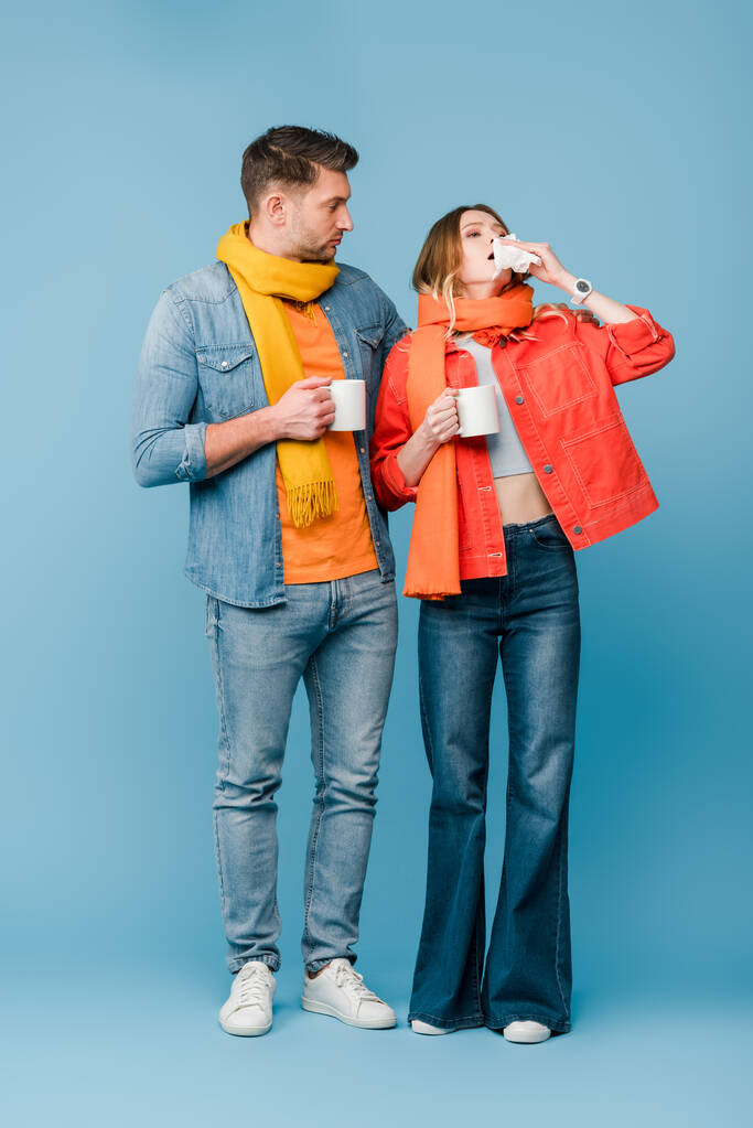 coppia malata in sciarpe in possesso di tazze con bevande calde mentre la donna starnutisce sul blu
 - Foto, immagini