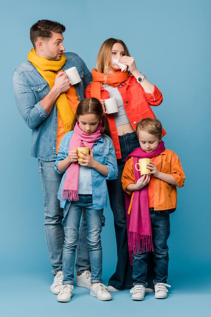λυπημένη άρρωστη οικογένεια με παιδιά σε κασκόλ κρατώντας κύπελλα με ζεστά ροφήματα στο μπλε - Φωτογραφία, εικόνα