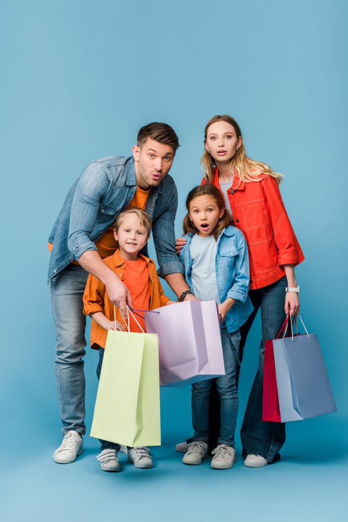 ενθουσιασμένοι γονείς και παιδιά έκπληξη κρατώντας τσάντες για ψώνια σε μπλε  - Φωτογραφία, εικόνα