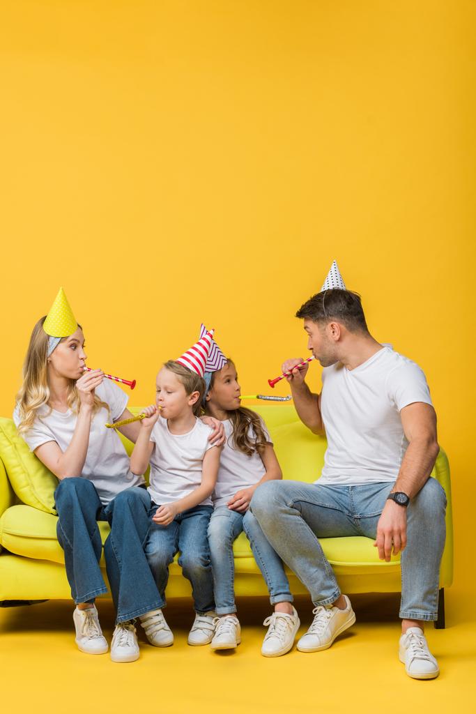 χαρούμενη οικογένεια στα γενέθλια καπάκια κόμμα με φυσητήρες στον καναπέ σε κίτρινο  - Φωτογραφία, εικόνα