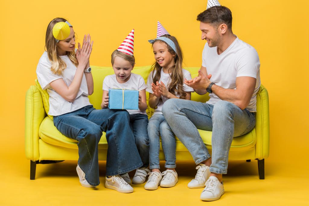 χαρούμενη οικογένεια σε καπελάκια πάρτι γενεθλίων, γιος κρατώντας κουτί δώρου στον καναπέ στο κίτρινο  - Φωτογραφία, εικόνα