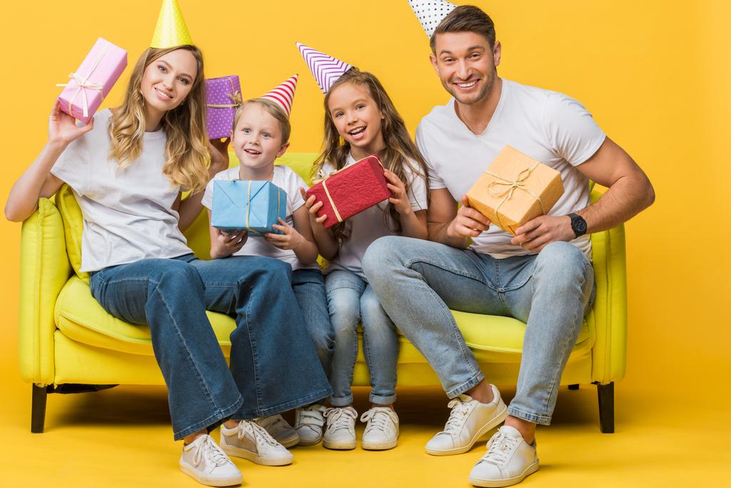 iloinen vanhemmat ja lapset syntymäpäiväjuhlissa lakit tilalla lahjapakkaukset sohvalla keltaisella
  - Valokuva, kuva