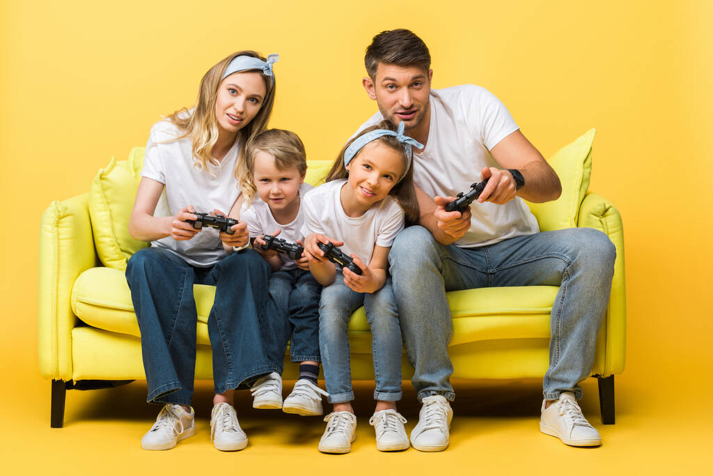 KYIV, UKRAINE - 4 MARS 2020 : joyeuse famille jouant à un jeu vidéo avec des joysticks assis sur un canapé jaune
 - Photo, image