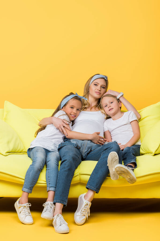beautiful pregnant mother hugging with daughter and son on sofa on yellow - Photo, Image