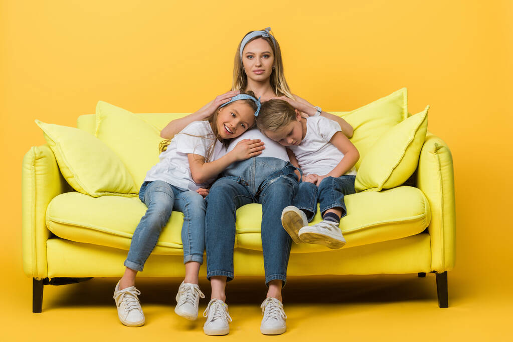 embarazada madre abrazo con sonriente hija e hijo en sofá en amarillo
 - Foto, imagen