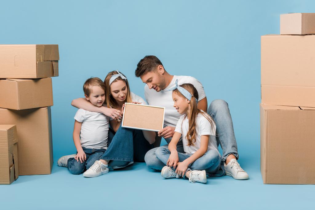 positive parents and kids holding frame and sitting on blue with cardboard boxes for relocation - Photo, Image