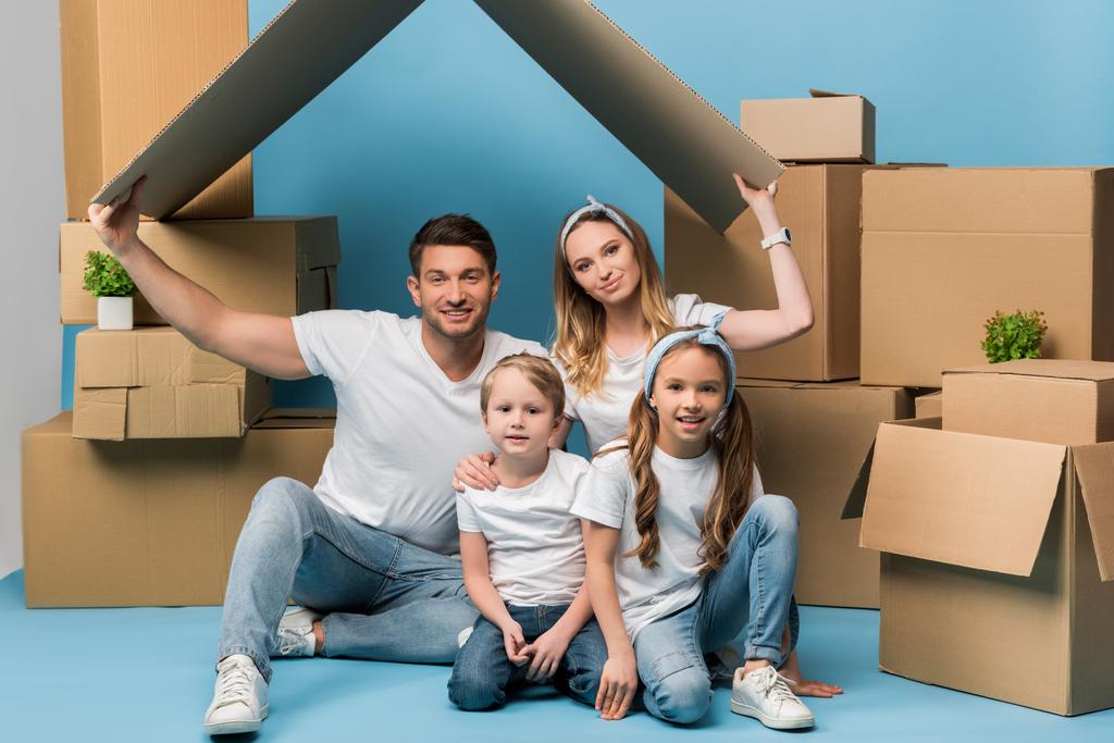 усміхнені батьки тримають картонний дах над дітьми на синьому з картонними коробками для переміщення
 - Фото, зображення