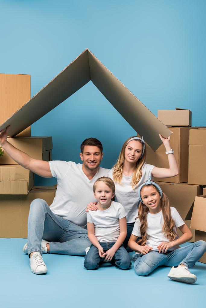 pozitivní rodiče drží lepenkovou střechu nad dětmi na modré s lepenkovými krabicemi pro relokaci - Fotografie, Obrázek