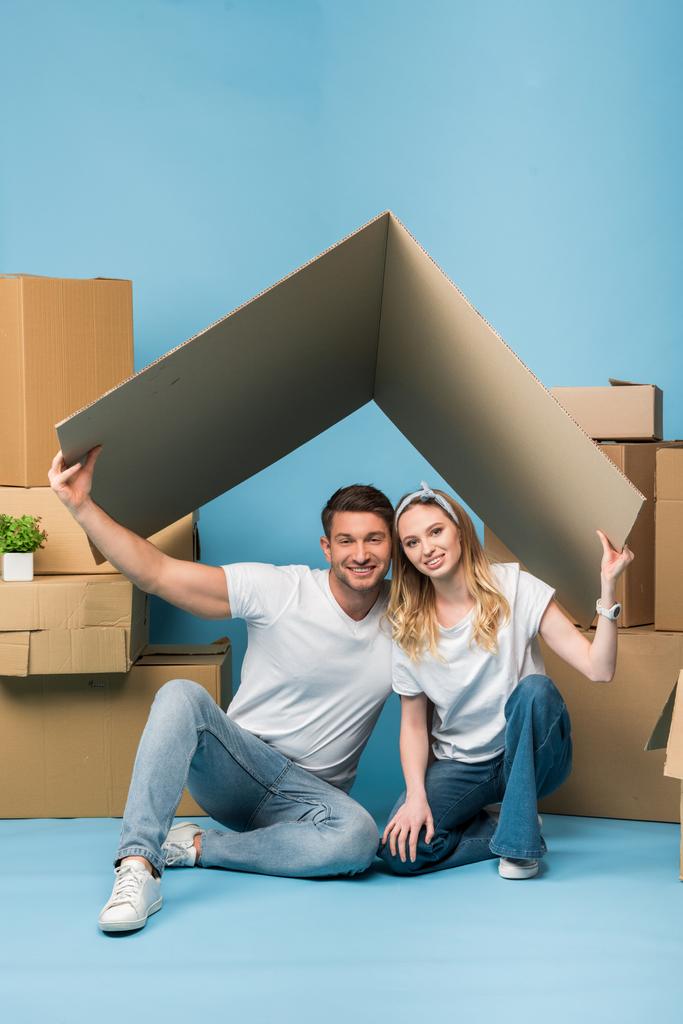 alegre pareja sosteniendo techo de cartón sobre cabezas mientras está sentado en azul con cajas de cartón para la reubicación
 - Foto, imagen