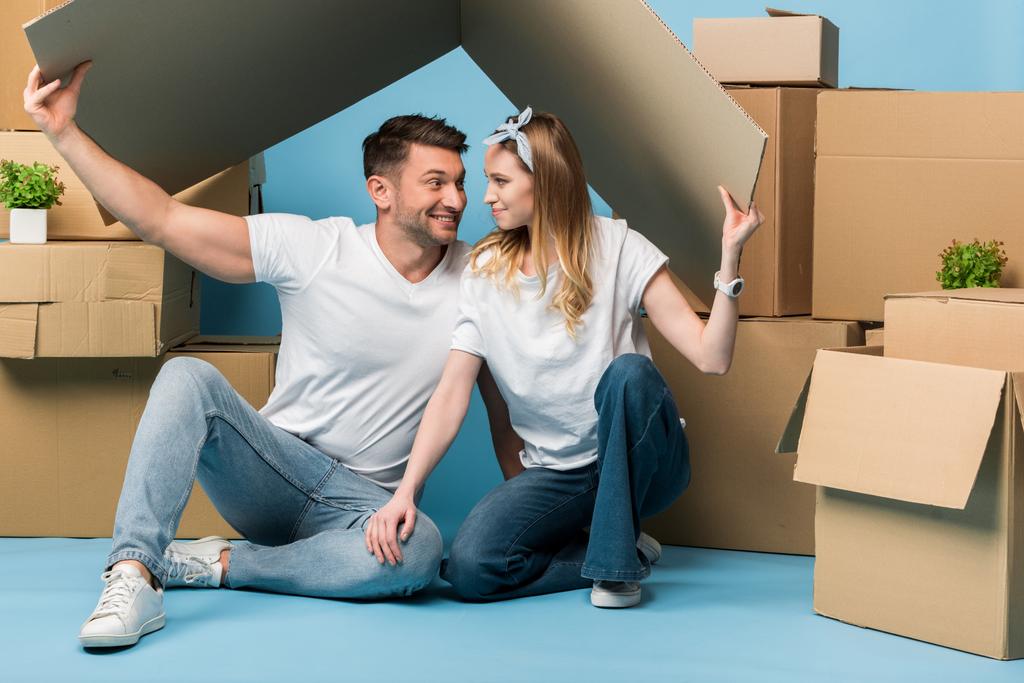 happy couple holding carton roof over heads while sitting on blue with cardboard boxes for relocation - Photo, Image