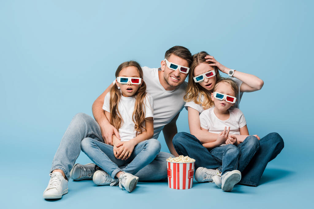 σοκαρισμένη οικογένεια σε 3d ποτήρια βλέποντας ταινία και κρατώντας τον κουβά ποπ κορν στο μπλε - Φωτογραφία, εικόνα