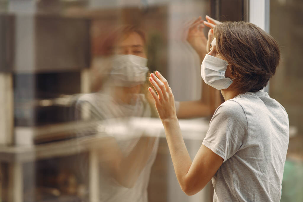 Γυναίκα με μάσκα στέκεται δίπλα στο παράθυρο. - Φωτογραφία, εικόνα