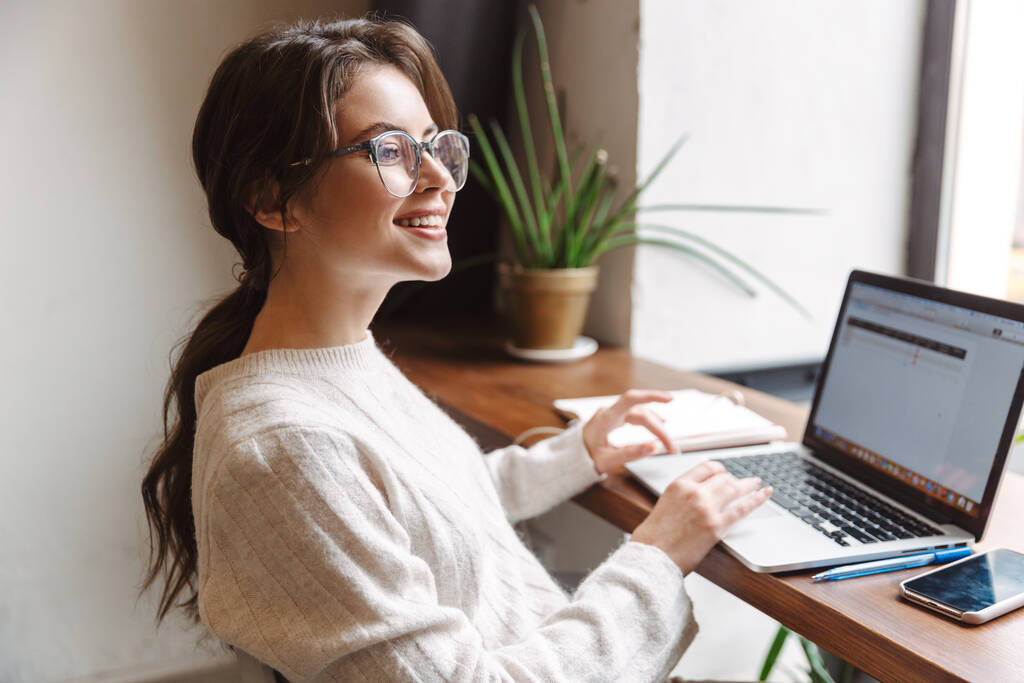 Kép fiatal gyönyörű fehér nő visel szemüveg segítségével laptop, miközben tanul a kávézóban - Fotó, kép