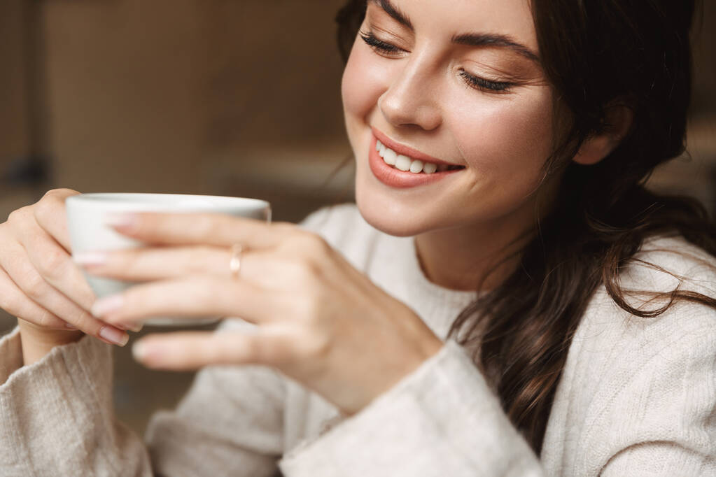 Изображение молодой красивой белой женщины с длинными каштановыми волосами, улыбающейся во время питья кофе в кафе
 - Фото, изображение