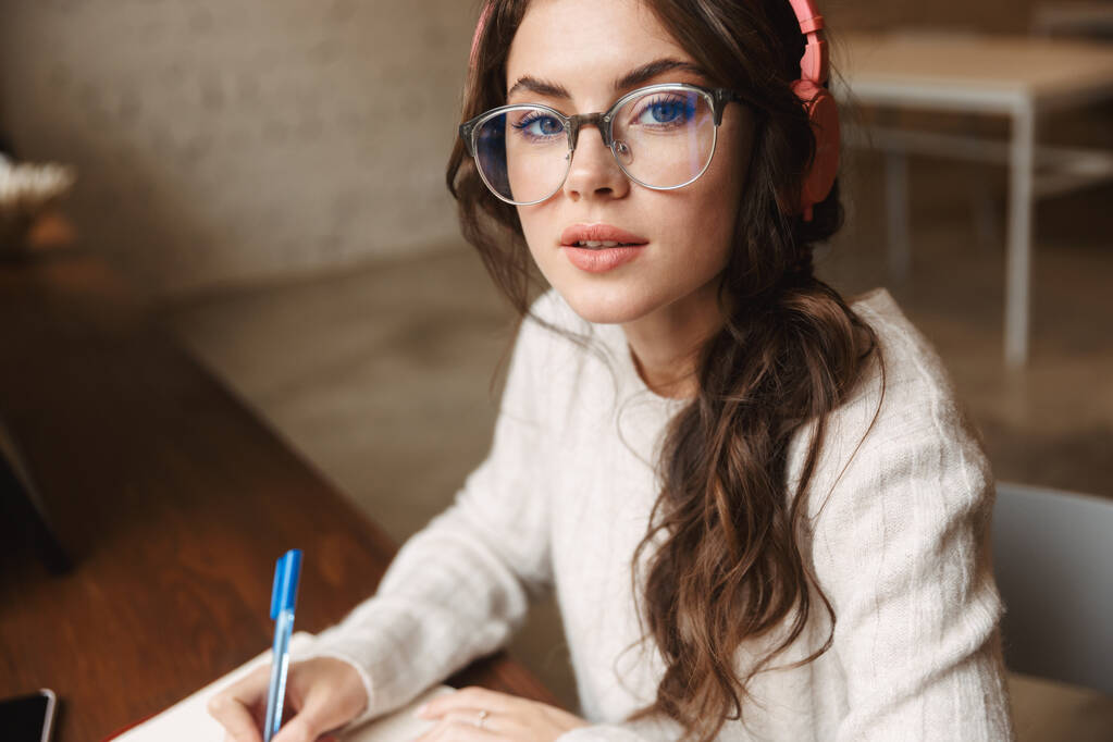 Imagem da jovem mulher branca bonita usando fones de ouvido estudando com caderno de exercícios no café
 - Foto, Imagem
