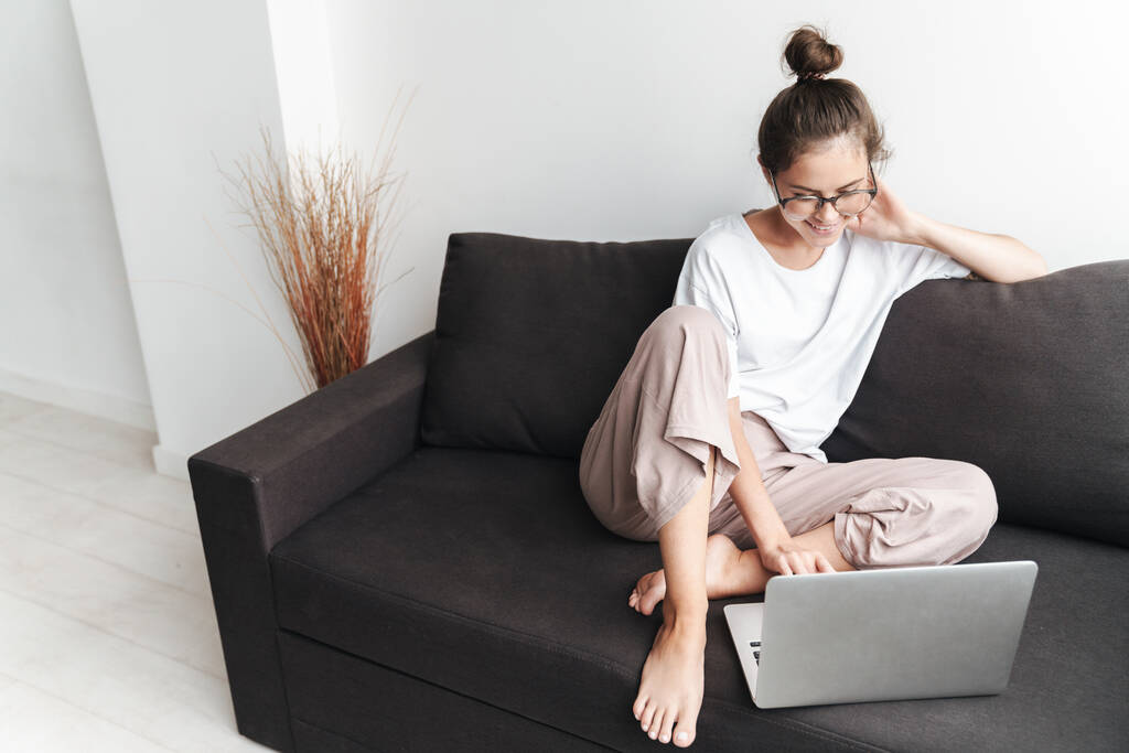 Bild einer lächelnden brünetten Frau mit Brille, die auf dem Laptop tippt, während sie im Wohnzimmer auf dem Sofa sitzt - Foto, Bild