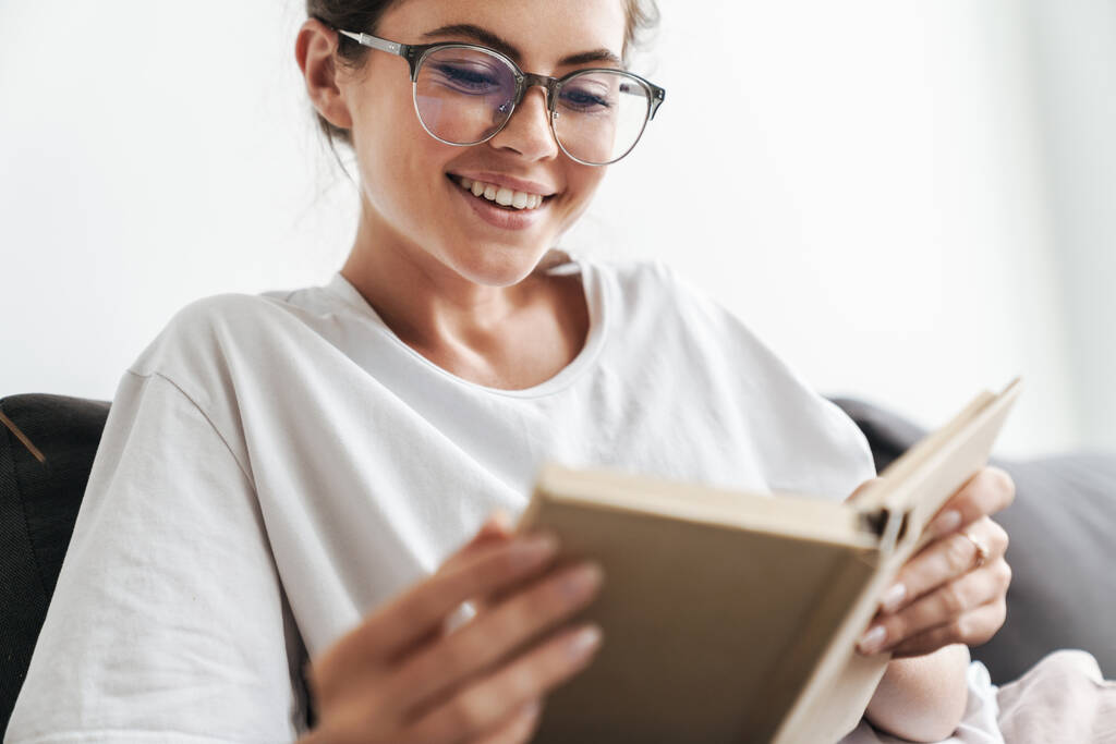 イメージクローズアップの喜びの白人女性で眼鏡をかけ本を読んでいる間にソファの上にリビングルームに座って - 写真・画像