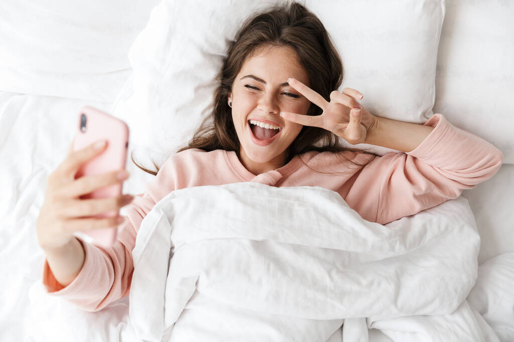 Top näkymä iloinen ihana nuori tyttö yllään pyjama makaa sängyssä makuuhuoneessa, ottaen selfie
 - Valokuva, kuva