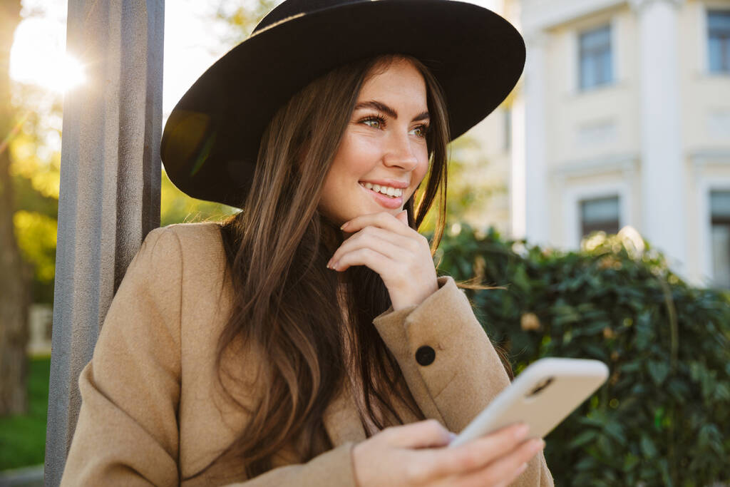 Ritratto di donna soddisfatta in cappotto e cappello autunnale che tiene lo smartphone mentre cammina all'aperto
 - Foto, immagini