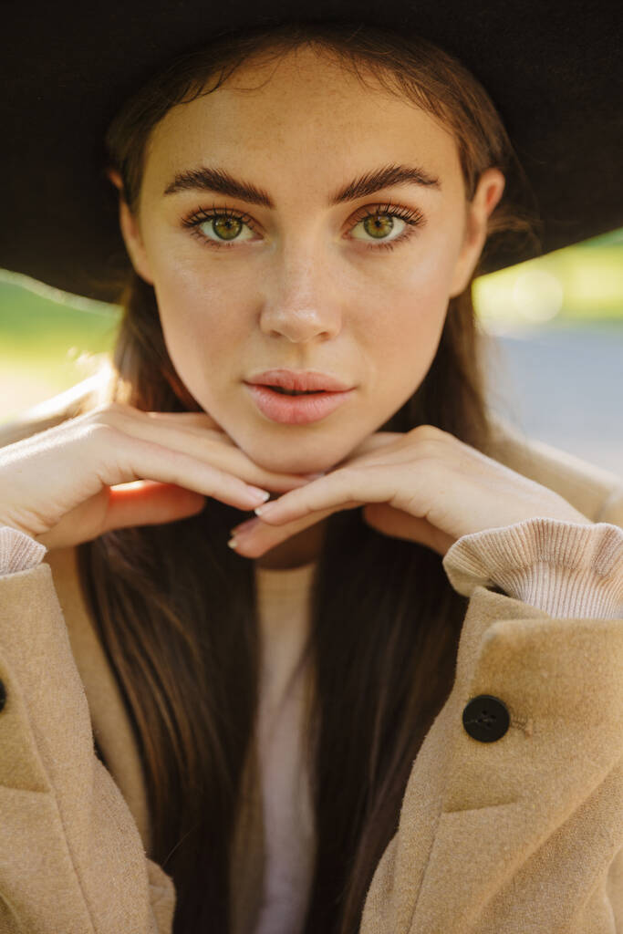 秋のコートを着た豪華な女性のイメージと、屋外の緑の公園でカメラを見る帽子 - 写真・画像