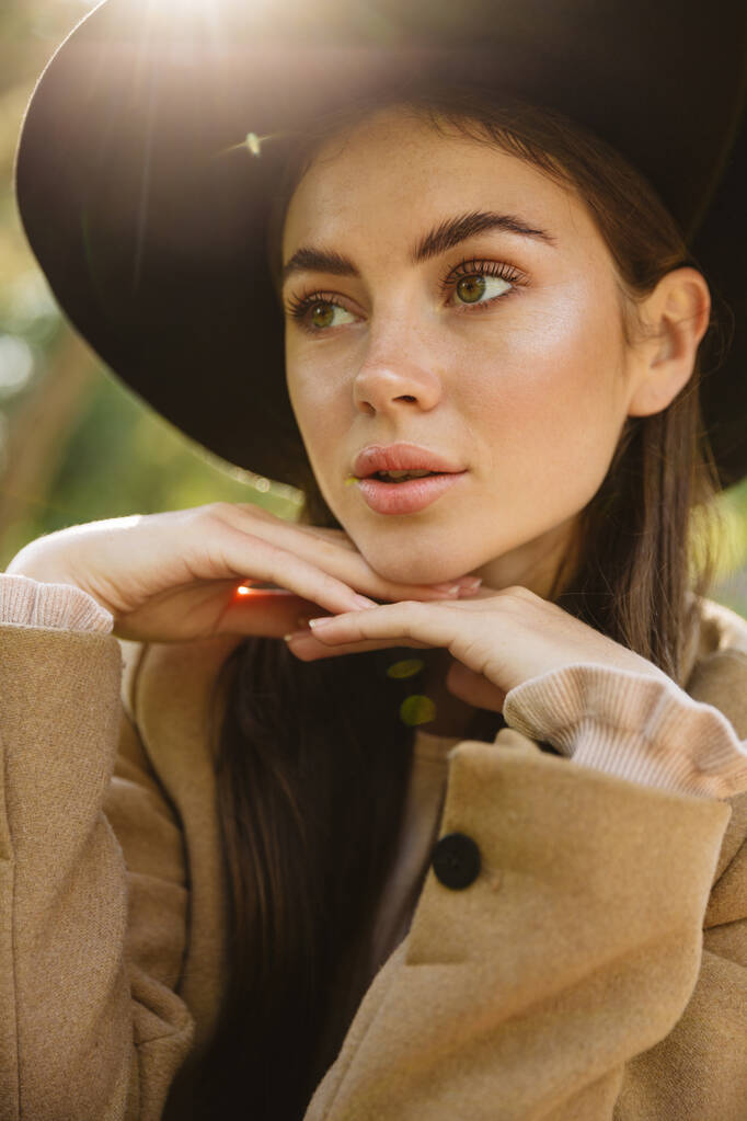Imagen de mujer adorable en abrigo de otoño y sombrero mirando a un lado en el parque verde al aire libre
 - Foto, imagen