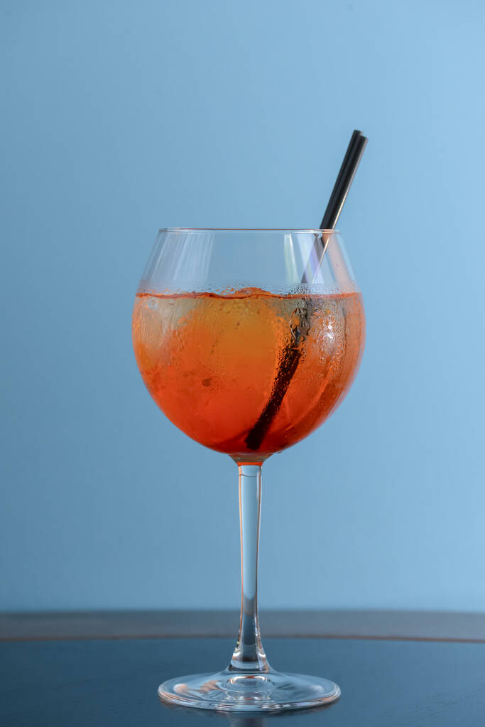 Coquetel de álcool frio de verão com gelo e laranja em um copo de vinho e palha de plástico preto no fundo azul pastel claro
 - Foto, Imagem