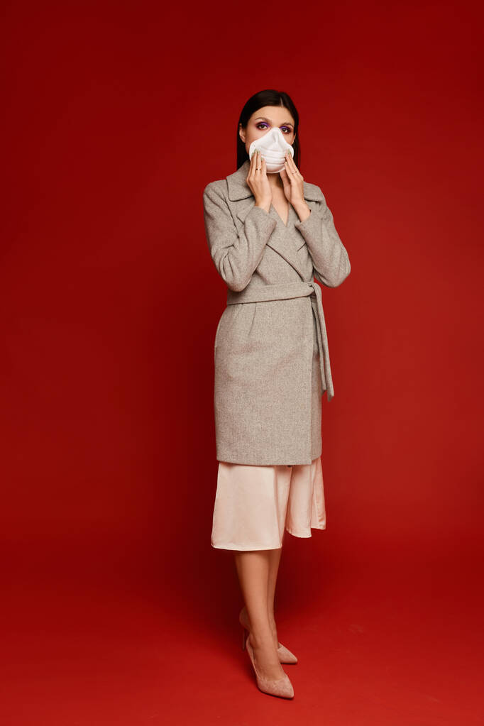 Piękna młoda kobieta w płaszczu i respiratorze pozująca na czerwonym tle z odizolowaną przestrzenią do kopiowania. Model dziewczyna w masce ochronnej i strój sprężynowy. Koncepcja zanieczyszczenia powietrza i grypy sezonowej - Zdjęcie, obraz
