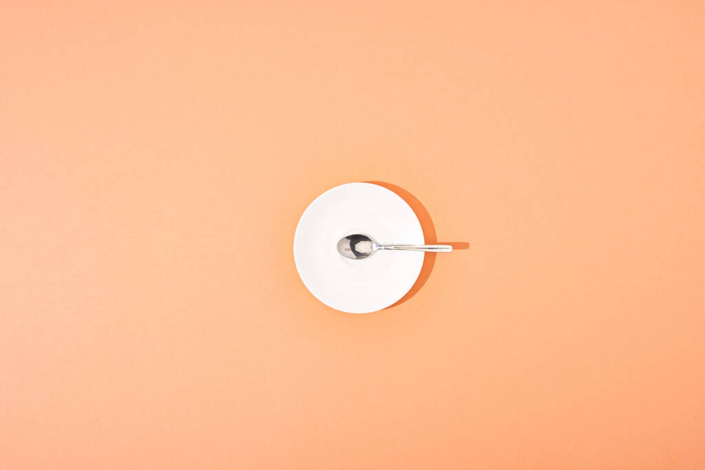 オレンジの背景にある白い丸皿の上からの金属スプーンの眺め - 写真・画像
