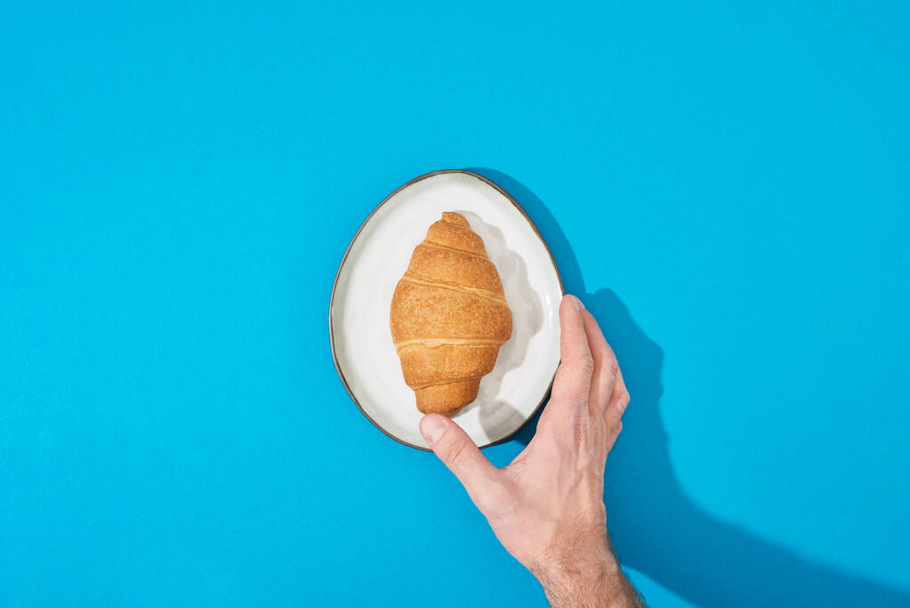 vista recortada del hombre sosteniendo croissant fresco en el plato sobre fondo azul
 - Foto, imagen