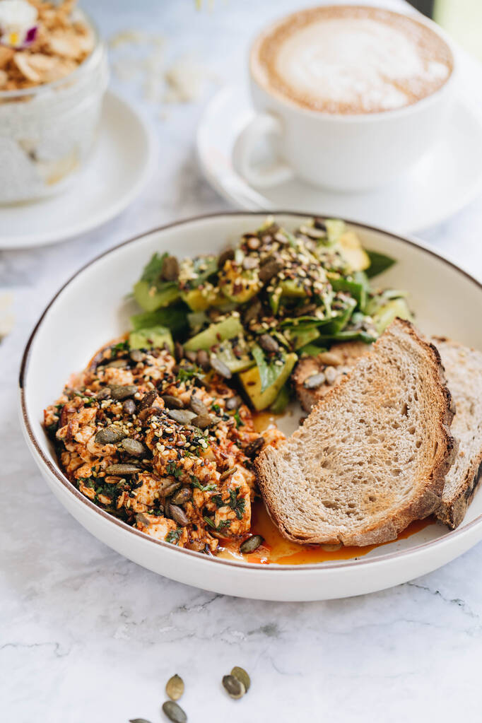 Tofu-sekoitus kurpitsansiemenillä, avokado-salaatti kurkulla ja basilikalla tarjoillaan rukiin paahtoleipää valkoisella levyllä marmoripohjalla
 - Valokuva, kuva