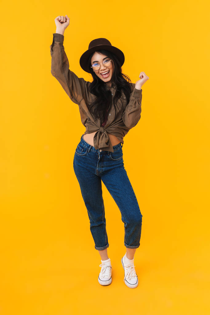 Full length image of young brunette asian woman wearing hat smiling and having fun isolated over yellow background - Photo, Image