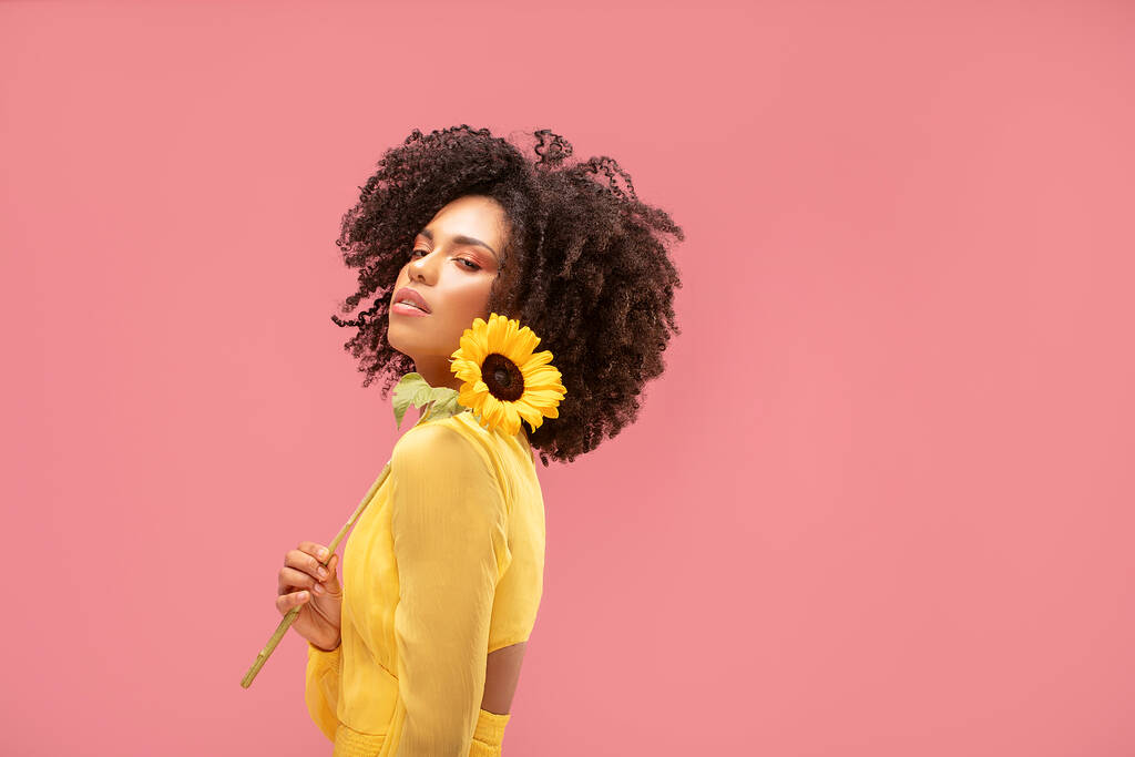 Pembe pastel stüdyo arka planında sarı ayçiçeğiyle poz veren güzel afro kadın. Kadınlar ve anneler günü kavramı. Bahar, yaz hisleri. Kız gücü. - Fotoğraf, Görsel