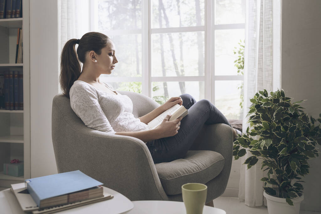 Όμορφη νεαρή γυναίκα χαλαρώνοντας στο σπίτι, είναι καθισμένος δίπλα σε ένα παράθυρο και διαβάζοντας ένα βιβλίο - Φωτογραφία, εικόνα