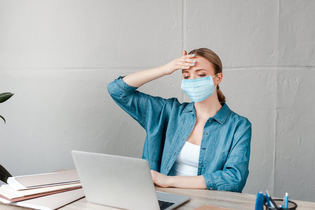 mulher em máscara médica tocando na testa enquanto trabalhava com laptop no escritório
 - Foto, Imagem