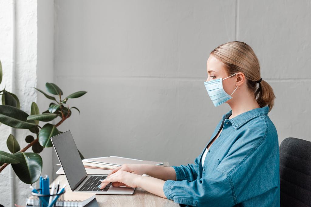 visão lateral da mulher em máscara médica trabalhando com laptop no escritório
 - Foto, Imagem
