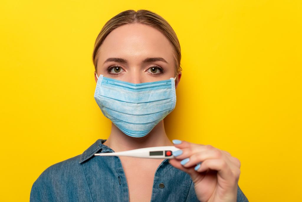 mujer joven en máscara médica sosteniendo termómetro sobre fondo amarillo
 - Foto, imagen