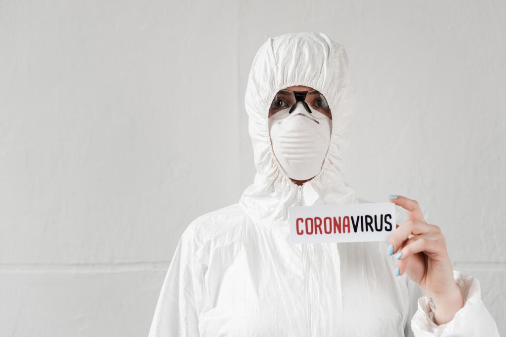 person in white hazmat suit, respirator and goggles holding card with coronavirus lettering - Photo, Image
