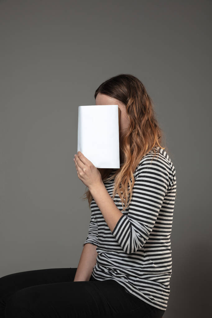 Feliz día mundial del libro, lee para convertirte en otra persona - mujer cubriendo la cara con un libro mientras lee sobre un fondo gris
 - Foto, Imagen