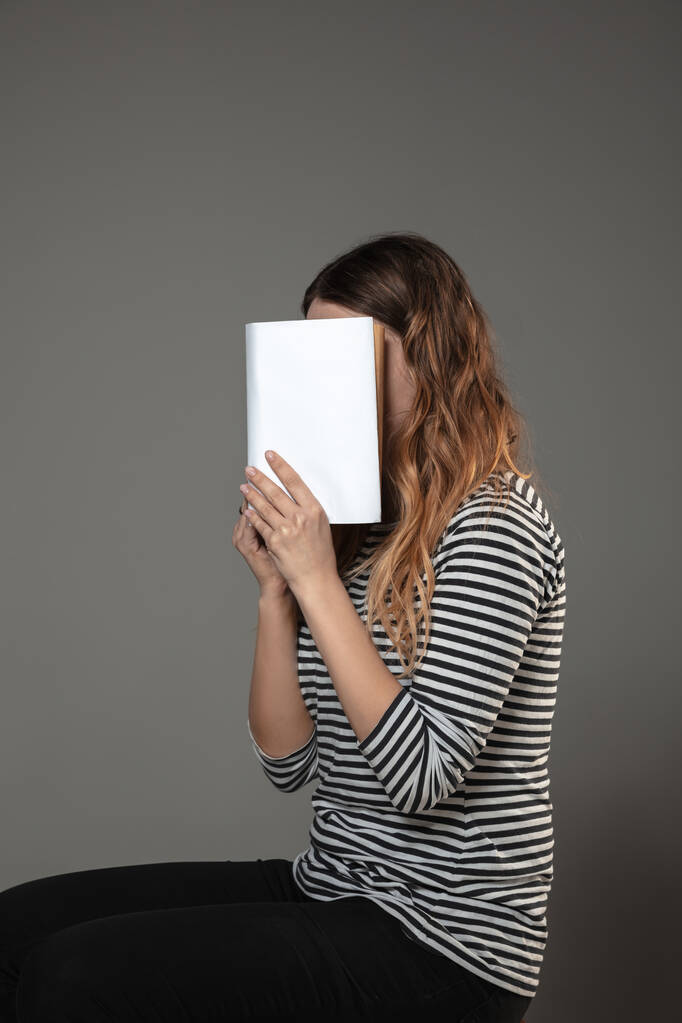 Feliz día mundial del libro, lee para convertirte en otra persona - mujer cubriendo la cara con un libro mientras lee sobre un fondo gris
 - Foto, Imagen